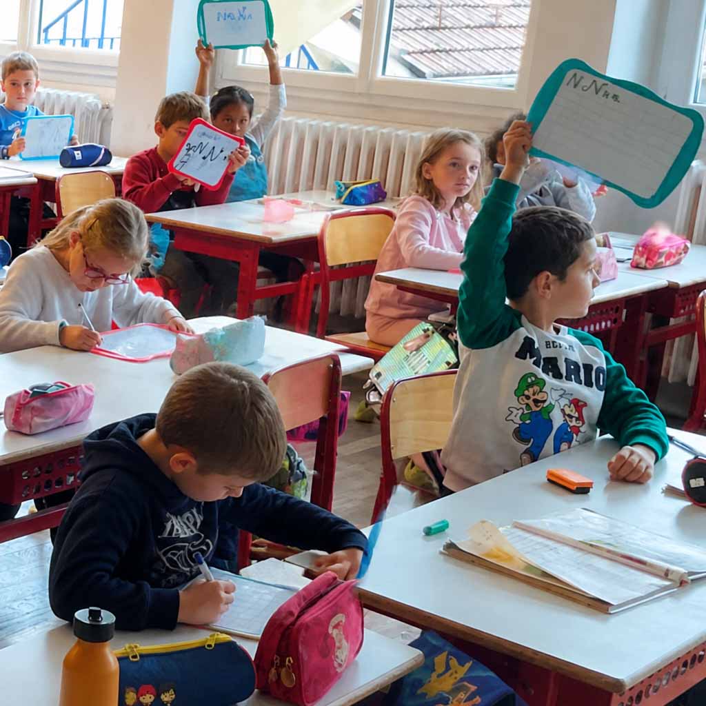 élèves de cp dans leur classe. Il lève leurs ardoises pour montrer la réponse